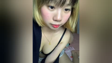A close-up video of an East Asian woman with shoulder-length blonde hair and fair skin, wearing a black bra and a necklace, sitting on a bed with a pink blanket.