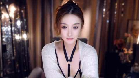 Video of an East Asian woman with light skin, brown hair in a bun, wearing a black harness and a white, ribbed cardigan, in a dimly lit, elegantly decorated room with a chandelier and curtains.