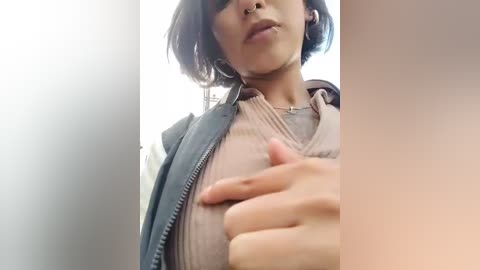 Video of a young woman with medium skin tone, short dark hair, and a septum piercing, wearing a grey jacket and a light grey shirt, standing in a bright room with soft, diffused light.