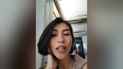 Media: Video of an Asian woman with straight black hair, wearing a beige top, standing in a dimly lit, narrow hallway. She has a nose ring and appears to be mid-conversation.