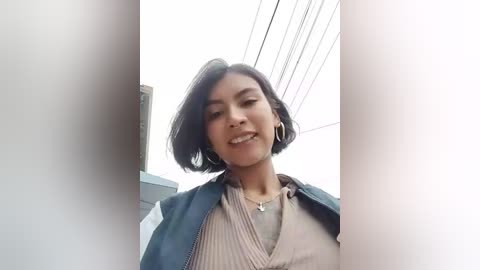 Video of a young woman with medium skin tone and short black hair, wearing a blue vest over a beige sweater, smiling. Background shows a white wall and power lines.