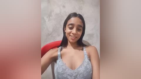 A video of a young woman with medium brown skin, long black hair, and a slender physique, wearing a light blue lace bralette, seated against a textured, light gray wall.