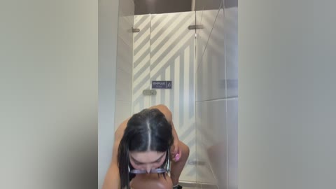 Video of a young woman with long black hair, wearing glasses, performing oral sex on a man. They are in a modern bathroom with white tiles and a grey door.