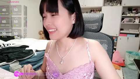 Video of an Asian woman with short black hair and a pale complexion, wearing a pink lace bra and pearl necklace, smiling while sitting on a bed with scattered clothes, against a cluttered background.