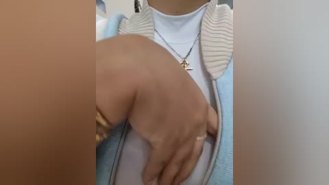 Video of a person with light skin, wearing a light blue zip-up jacket over a white shirt. They have a silver necklace with a star pendant and are gently caressing their abdomen. The background is blurred, suggesting an indoor setting.