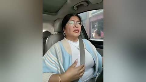 Video of a woman in a car, wearing glasses, light sweater, and a seatbelt, with a blurred background.