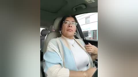 Video of a woman in a car, wearing a light blue and white sweater over a white shirt, and clear glasses. She has medium-length dark hair and is adjusting her sweater.