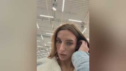 Video of a young, light-skinned woman with straight brown hair, wearing a light blue denim jacket, looking introspective in an industrial warehouse setting with exposed pipes and fluorescent lights.