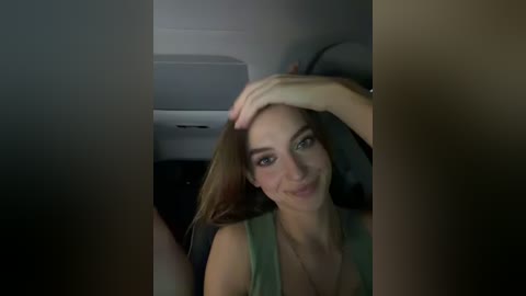 Video of a smiling woman with long, light brown hair, wearing a sleeveless green top, sitting in a dimly lit car. Her hand rests on her head.