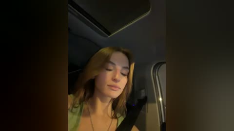 A video captures a young woman with long, light brown hair, wearing a green top, leaning back in a car seat with eyes closed. The dim interior lighting casts a warm, ambient glow.