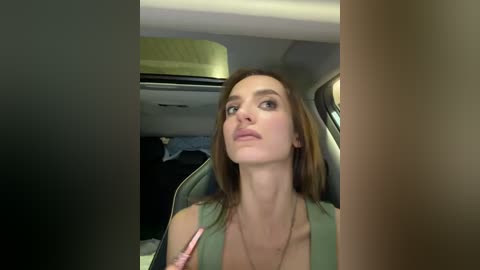 Video of a Caucasian woman with straight brown hair and light skin, wearing a green top, holding a pink pen, seated in a car with beige seats and a visible overhead light.