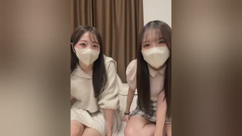 Video of two young East Asian women with long black hair, wearing beige face masks and cream sweaters, sitting on a beige floor in front of brown curtains.