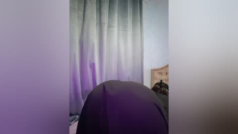 Video of a dimly lit bedroom with a person lying face-down on a bed, covered in a dark blanket. A light gray curtain with a soft purple hue partially obscures the window, casting a subtle glow.