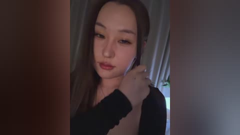 A video of an East Asian woman with fair skin, long brown hair, and light makeup, wearing a black top, adjusting her earphones. The background features beige curtains and a potted plant.