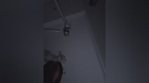 Video of a dimly lit room with a ceiling-mounted microphone capturing a shadowy figure in the corner. The room's walls are a muted gray, and the lighting is low, creating a moody atmosphere.