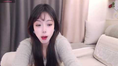 Video of an Asian woman with long black hair, wearing a grey cardigan, sitting on a beige couch, with a shocked expression, surrounded by beige curtains.