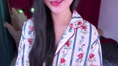 Video of an Asian woman with long black hair, wearing a floral-patterned white shirt, smiling. Background shows a plush toy and red curtains.