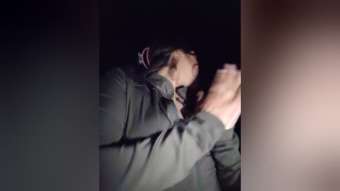 Media: Video of a young woman with long black hair, wearing a dark jacket, looking distressed, possibly in pain, with a blurred, dimly lit background.