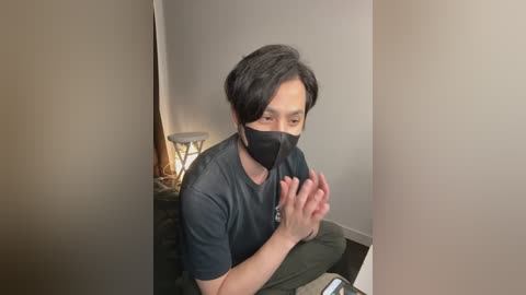 A video of a young Asian man with short black hair, wearing a black face mask and dark t-shirt, sitting on a couch, clapping hands in a small, dimly lit room.