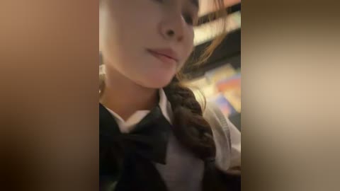 Video of a close-up of a young woman with fair skin, brown hair, and a soft smile, wearing a white blouse and black bow tie, against a blurred background.