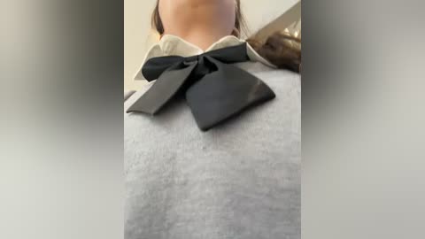 A close-up video of a person wearing a gray sweater with a large, black bow at the collar, partially obscured by a blurred background.