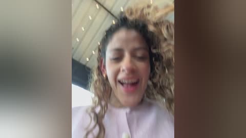 Media: Video of a curly-haired, light-skinned woman with a joyful expression, wearing a light pink shirt, against a blurred indoor setting with wooden ceiling beams.
