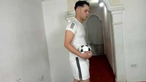 Video of a fit, fair-skinned man with short black hair in a white athletic outfit holding a black-and-white soccer ball in a narrow, dimly lit hallway with white walls and red carpet.