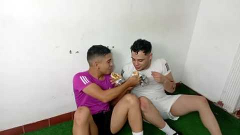 Video of two young men, one in a purple shirt, sitting on a green mat, sharing a sandwich in a white-walled room with red tiles.