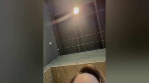 A video shows a woman's face, looking down, with her hair tied back, in a dimly lit room with a wooden ceiling and a single, bright light bulb overhead.