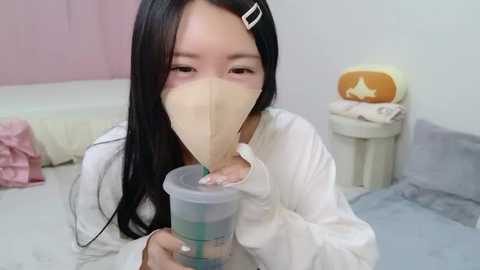 A video of an East Asian woman with long black hair, wearing a beige face mask and white long-sleeve shirt, drinking from a clear bottle with a straw in a modestly furnished room with pink walls and a white bed.