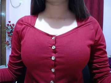 Video of a woman with medium skin tone, wearing a red button-up cardigan with long sleeves. She has medium-sized breasts, dark brown hair, and is indoors with a white wall and red curtains in the background.