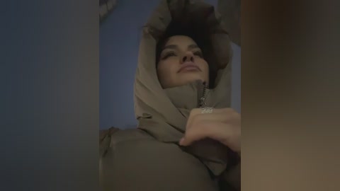 Video of a woman with medium skin tone, wearing a beige coat, her eyes closed, and arms raised, against a dimly lit, blue background.