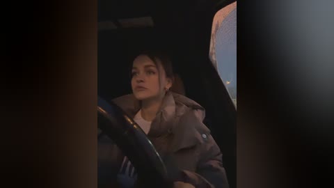 Media: A dimly-lit video of a young woman in a car, wearing a beige coat, with her hair tied back, focused on driving, partially obscured by a dark shadow.