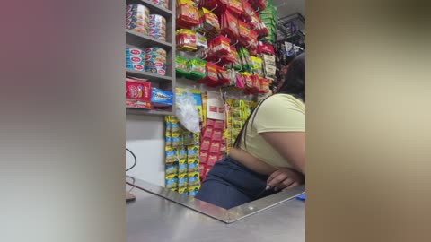 Video of a female cashier with long hair, wearing a yellow shirt and dark pants, standing behind a glass counter, surrounded by colorful snack bags.
