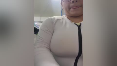 Video of a light-skinned Asian woman with glasses, wearing a tight, white long-sleeved shirt with a black zipper, smiling slightly, partially obscured by a frosted glass door. Background shows a cluttered room with shelves.