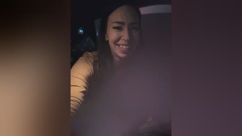 Video of a smiling, fair-skinned woman with straight, dark hair in a dimly lit car, wearing a beige top.