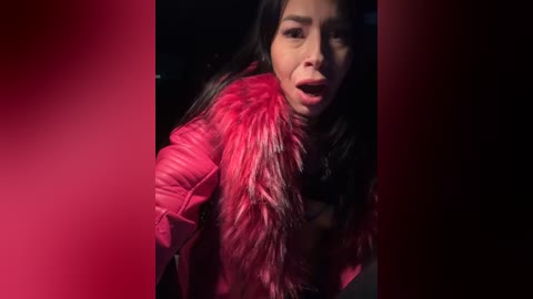 Video of a woman with long black hair, wearing a bright pink fur jacket, looking shocked or surprised, with a dark background.