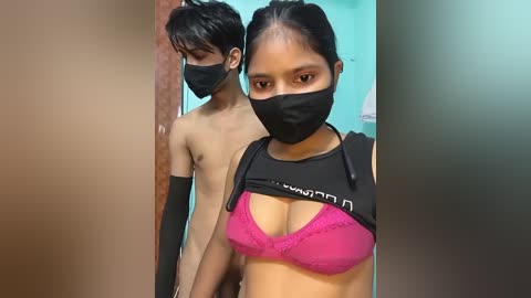 Video of two shirtless Asian men, one with a black mask, and a woman with a black mask, pink bra, and black gloves, in a teal room.