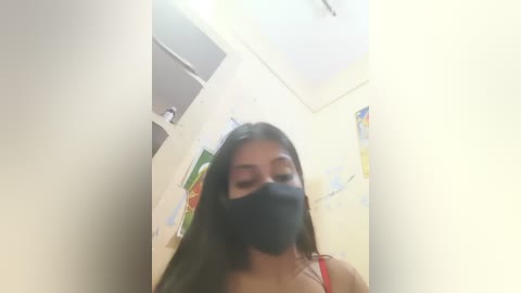 A video of a young woman with long black hair, wearing a black face mask, a red tank top, and standing in a cluttered, brightly lit room with posters on the walls.