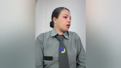 Video of a Latina woman with medium-dark skin, dark hair in a bun, wearing a gray uniform with a black tie featuring a green and blue logo. She looks serious.