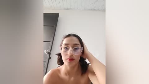 Video of a young Asian woman with shoulder-length dark hair, fair skin, and large, clear-framed glasses, standing topless in a modern, minimalist room with white walls and a dark door.