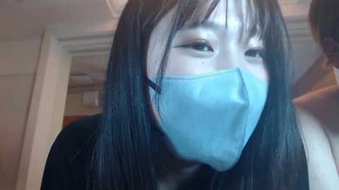 Video of a young Asian woman with long black hair and pale skin, wearing a blue medical mask, looking downward, indoors with a blurred background.
