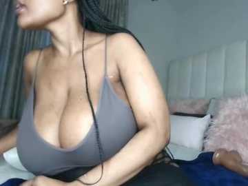 Video of a light-skinned woman with long black braids, wearing a low-cut gray tank top, revealing ample cleavage, in a modern, minimalistic bedroom with white pillows and a white headboard.
