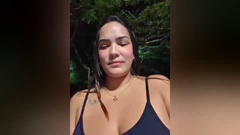 Video of a young woman with long black hair, medium skin tone, and a slender physique, wearing a black spaghetti-strap top, outdoors with blurred green foliage background.