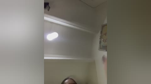 Video of a ceiling with a bright, rectangular light fixture. The walls are beige, and a framed picture hangs on the right. The image appears slightly blurry, possibly due to movement or low light.