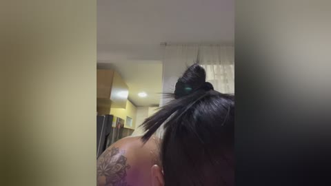Media: Video of a person's upper back and head with black hair in a bun, showcasing a large tattoo on the shoulder. Background features a modern, softly lit room with yellow walls and white curtains.