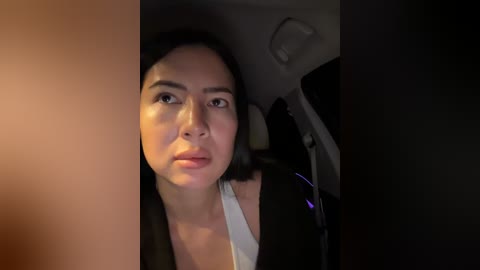 Video of a woman with straight black hair, fair skin, and light makeup, wearing a white tank top and a black jacket, seated in a dimly lit car, with a neutral expression.