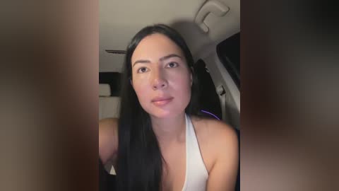 Media: Video of a young woman with long black hair, wearing a white tank top, taken from a car, showing the front seat and part of the dashboard.
