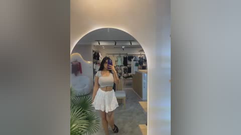 Video of a young woman with light brown skin and straight black hair, wearing a white crop top and high-waisted white skirt, taking a selfie in a mirrored hallway with soft lighting.
