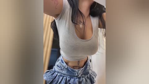 Media: Video of a woman with long, dark hair, wearing a tight grey crop top that accentuates her ample breasts, and high-waisted, frayed denim shorts, with a visible tattoo on her lower abdomen.